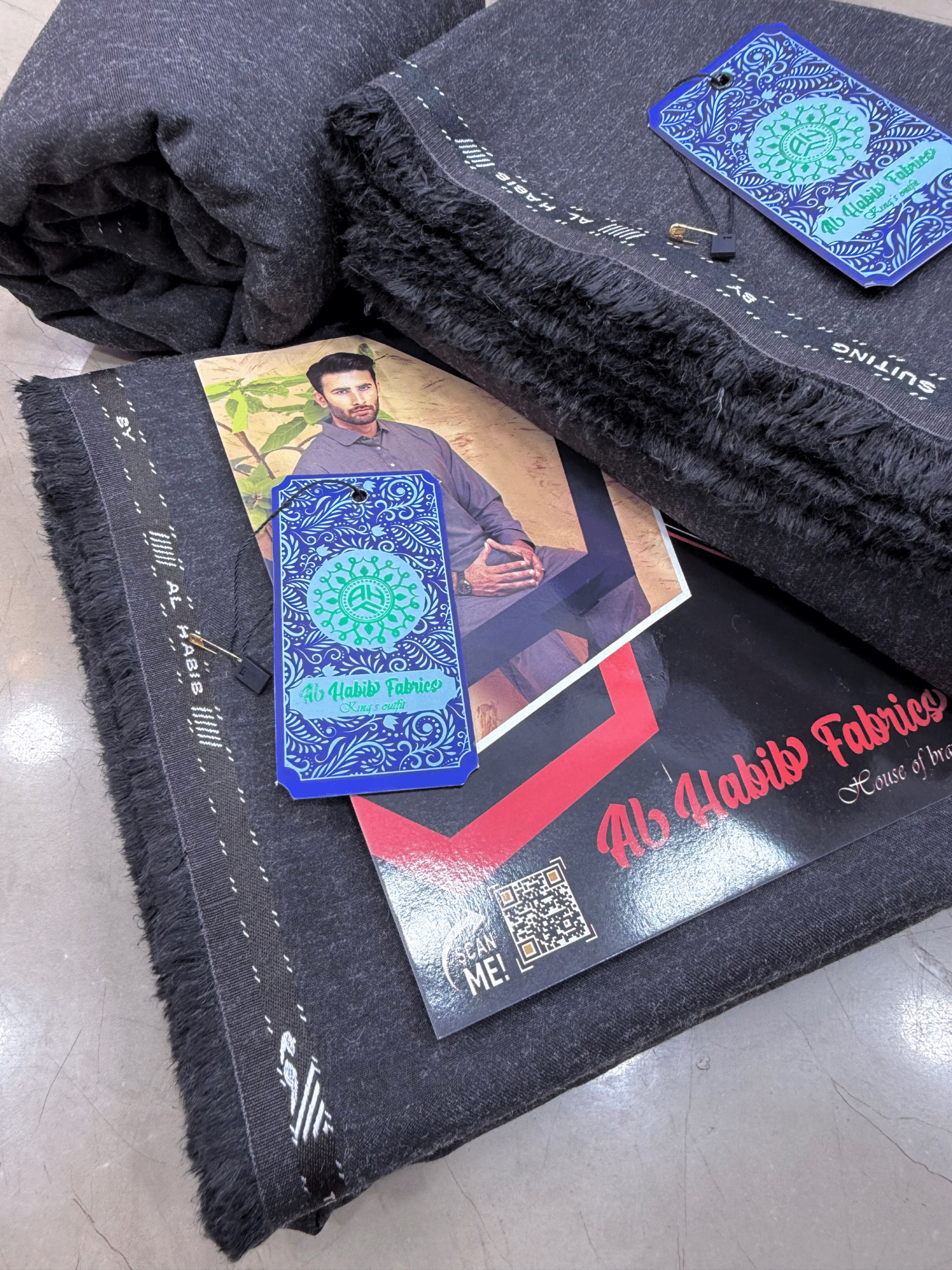 Black fabric with branded tags and a card featuring a person and brand **AlHabibFabrics, AlHabibWinter, King'sOutfit, TopClassChoice, AlHabib, KashmirWool, WoolFabric, PremiumWool, SoftWool, WarmFabric, UnstitchedWool, SuitingWool, Men'sSuiting, UnstitchedFabric, ShalwarKameezFabric, KurtaShalwarFabric, Men'sDressMaterial, PremiumSuit, LuxuryMenswear, ElegantFabric, FormalWearMen, BestWinterFabric, NavyBlue, CharcoalGrey, DarkBrown, DeepBlue, MaroonFabric, GreySuit, PlainColors, PakistaniFabric, Menswelogo.