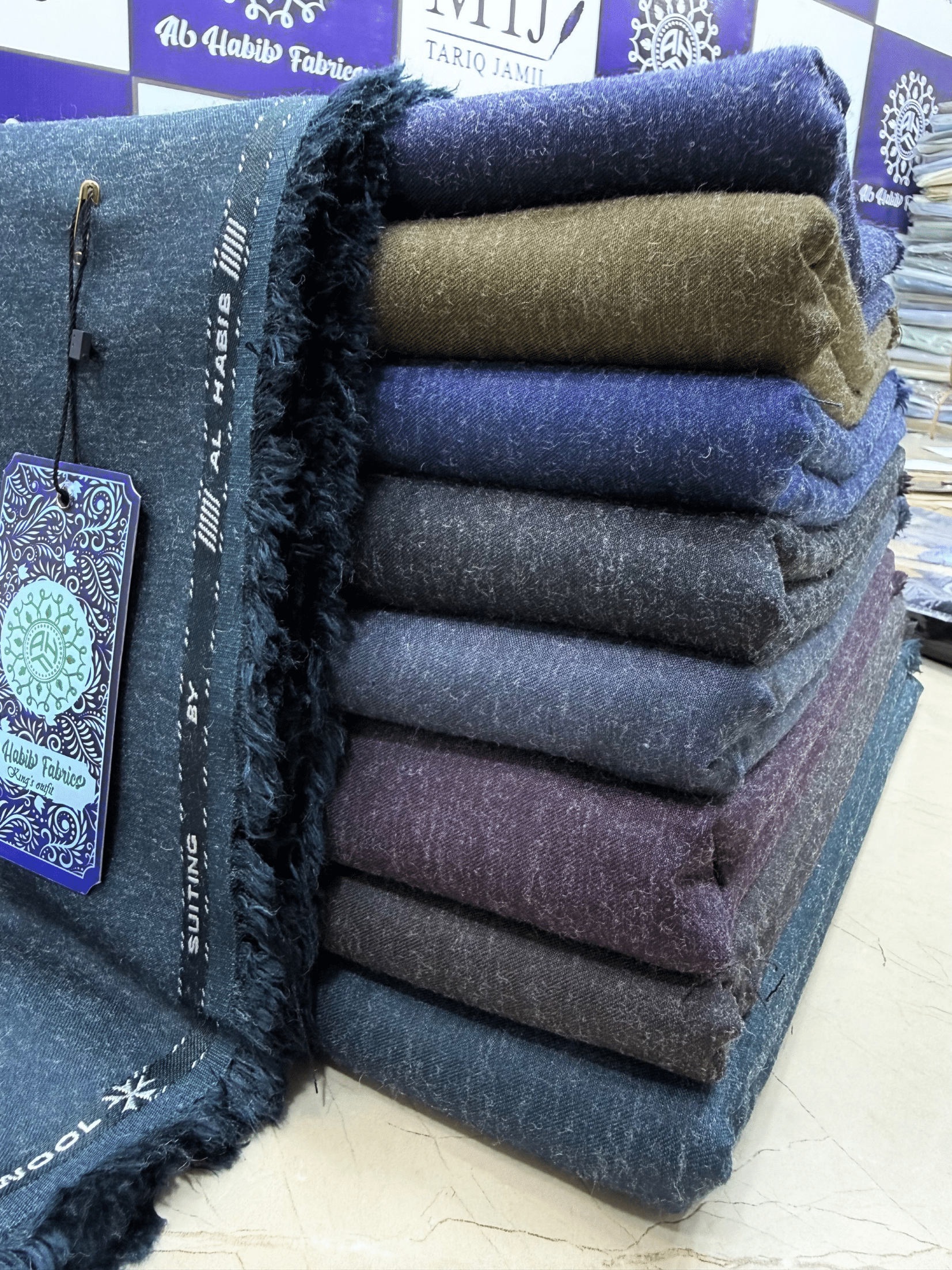 Stack of folded fabric swatches in various colors with a visible brand **AlHabibFabrics, AlHabibWinter, King'sOutfit, TopClassChoice, AlHabib, KashmirWool, WoolFabric, PremiumWool, SoftWool, WarmFabric, UnstitchedWool, SuitingWool, Men'sSuiting, UnstitchedFabric, ShalwarKameezFabric, KurtaShalwarFabric, Men'sDressMaterial, PremiumSuit, LuxuryMenswear, ElegantFabric, FormalWearMen, BestWinterFabric, NavyBlue, CharcoalGrey, DarkBrown, DeepBlue, MaroonFabric, GreySuit, PlainColors, PakistaniFabric, Mensweatag.