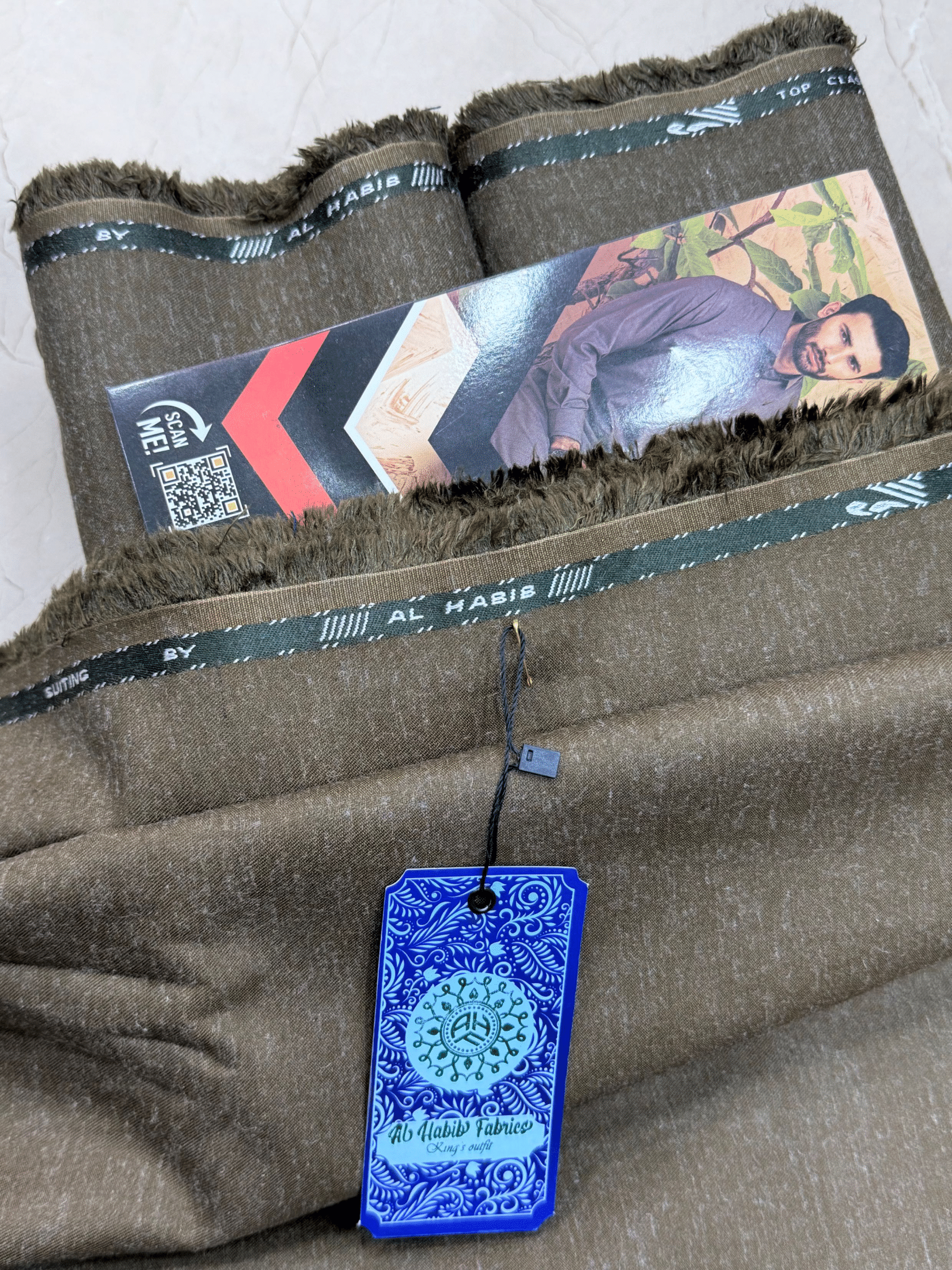 Folded fabric with a blue tag and visible branding on a light gray **AlHabibFabrics, AlHabibWinter, King'sOutfit, TopClassChoice, AlHabib, KashmirWool, WoolFabric, PremiumWool, SoftWool, WarmFabric, UnstitchedWool, SuitingWool, Men'sSuiting, UnstitchedFabric, ShalwarKameezFabric, KurtaShalwarFabric, Men'sDressMaterial, PremiumSuit, LuxuryMenswear, ElegantFabric, FormalWearMen, BestWinterFabric, NavyBlue, CharcoalGrey, DarkBrown, DeepBlue, MaroonFabric, GreySuit, PlainColors, PakistaniFabric, Menswbackground